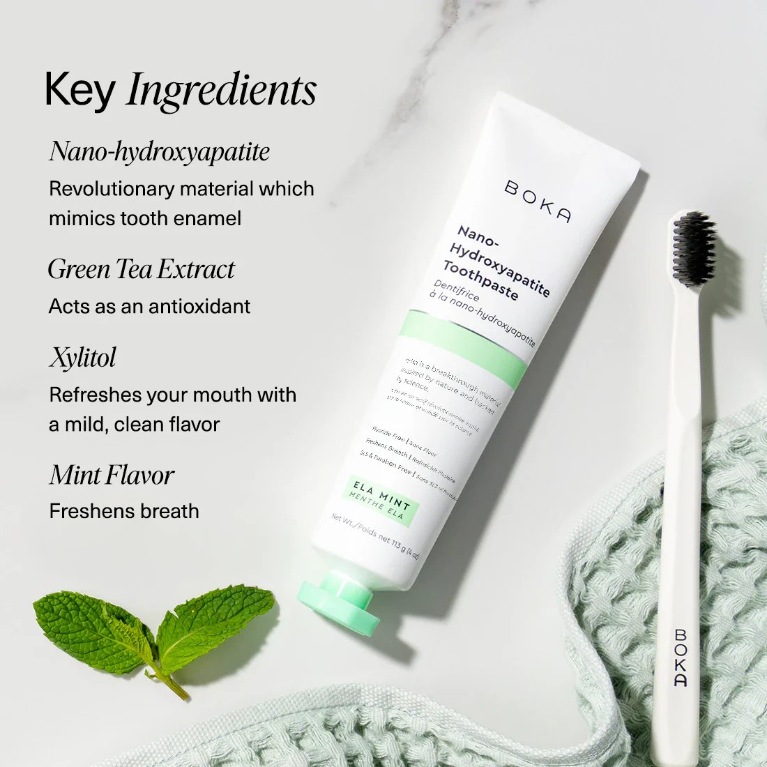 Boka Ela Mint toothpaste tube and toothbrush on a textured towel with mint leaves, alongside key ingredients listed